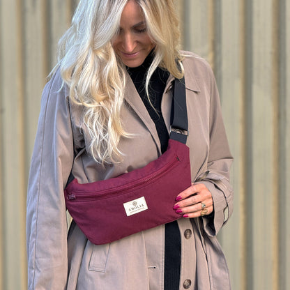 Woman wearing a beige trench coat holding a purple waist bag with a visible brand logo.